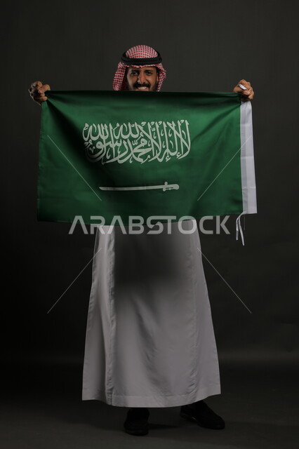 Standing straight and looking at the camera with joyful gestures on Flag Day, March 11, feelings of enthusiasm during the celebration of Saudi National Day, September 23, portrait of a Saudi Gulf Arab man wearing a shemagh and traditional thobe, holding the Kingdom’s flag in his hand, full-length body portrait, gray background