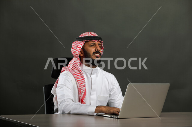 Advanced and modern means and technologies, a close-up portrait of a Saudi Gulf Arab man wearing traditional clothing and a shemagh, chatting via the laptop, receiving shocking news, completing office tasks via the laptop, gray background