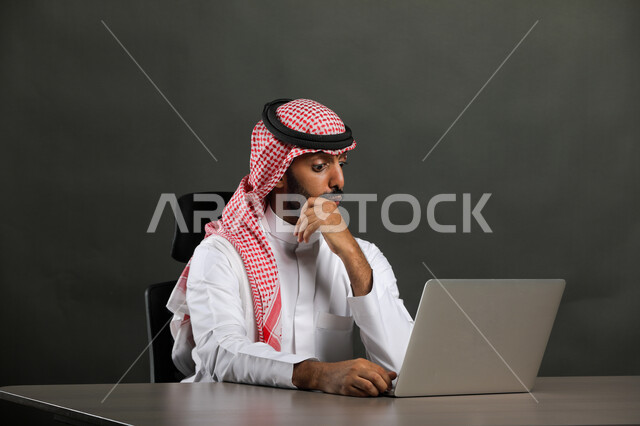 إنجاز العمل اونلاين من خلال الحاسب المحمول، بورتريه مقرب  لرجل عربي خليجي سعودي يرتدي الزي التقليدي والشماغ يجلس على المكتب ويستخدم جهاز اللابتوب، ايماءات التفكير والحيرة، خلفية رمادي