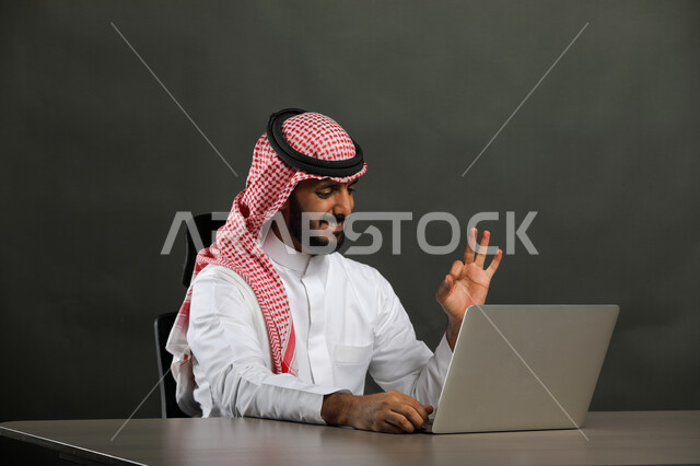 Completing office tasks via the laptop, a close-up portrait of a Saudi Gulf Arab man wearing a traditional dress and shemagh, sitting at the desk in front of the laptop, making successful deals with clients via the laptop, gestures of enthusiasm and happiness, gray background