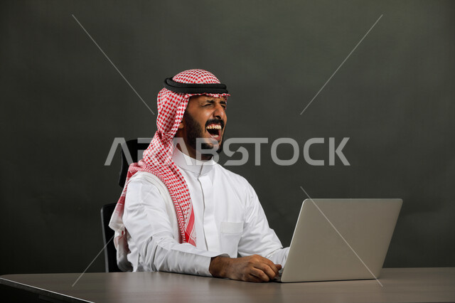 إستخدام جهاز الحاسوب المحمول، بورتريه مقرب  لرجل عربي خليجي سعودي يرتدي الزي التقليدي والشماغ و يقوم بالنظر الى شاشة الحاسوب المحمول، القيام بصفقات ناجحة مع العملاء عبر اللابتوب، ايماءات الضحك   والسعادة، خلفية رمادي