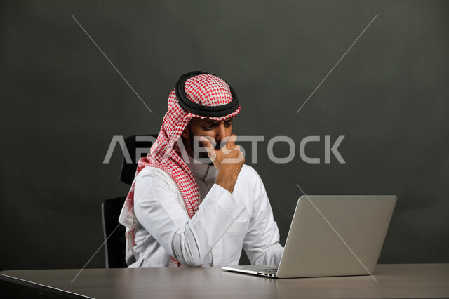  إحتراف وظائف الحاسوب وتكنولوجيا المعلومات، بورتريه مقرب  لرجل عربي خليجي سعودي يرتدي الزي التقليدي والشماغ و يجلس على المكتب أمام اللابتوب، ايماءات الحيرة والتفكير، إستخدام الأجهزة التقنية الحديثة، خلفية رمادي