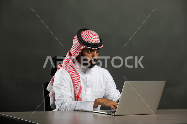 Using modern technologies in remote work, a close-up portrait of a Saudi Gulf Arab man wearing a traditional dress and shemagh, looking at the laptop screen, receiving shocking news over the Internet, doing work online through the laptop, gray background