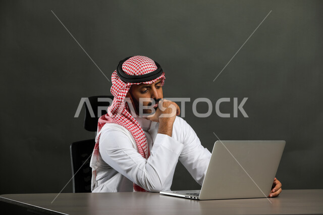 إنجاز العمل اونلاين من خلال الحاسب المحمول، بورتريه مقرب  لرجل عربي خليجي سعودي يرتدي الزي التقليدي والشماغ و يقوم بإجراء مكالمة فيديو، تطور التكنولوجيا وإستخدام الأجهزة التقنية الحديثة، ايماءات الحيرة والتفكير،  خلفية رمادي