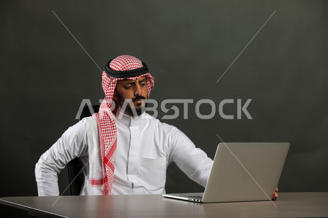 Using a laptop, a close-up portrait of a Saudi Gulf Arab man wearing a traditional dress and a shemagh, sitting at the desk and using the laptop to accomplish tasks, advanced and modern methods and technologies, watching movies and series online, gray background.