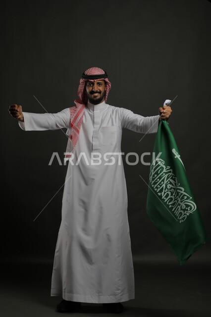إيماءات السعادة والحماس في الاحتفال باليوم الوطني السعودي 23 سبتمبر، بورتريه لرجل عربي خليجي سعودي يرتدي الشماغ والثوب التقليدي يحمل بيده راية المملكة ، النظر إلى الكاميرا بإيماءات الفوز والنصرفي يوم العلم  11 مارس ، صورة الجسم بالطول الكامل , خلفية رمادية