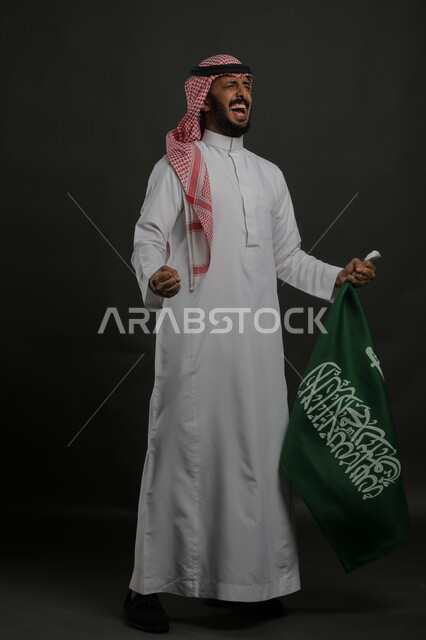 إيماءات الاحتفال باليوم الوطني السعودي 23 سبتمبر، بورتريه لرجل عربي خليجي سعودي يرتدي الشماغ والثوب التقليدي يحمل بيده علم المملكة ،الفرح بيوم العلم  11 مارس ، صورة الجسم بالطول الكامل , خلفية رمادية