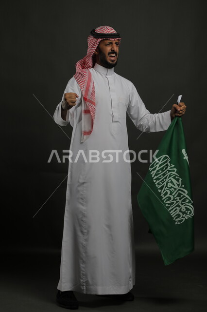 الشعور بالحماس أثناء الاحتفال باليوم الوطني السعودي 23 سبتمبر، بورتريه لرجل عربي خليجي سعودي يرتدي الشماغ والثوب التقليدي يحمل بيده علم المملكة ،إيماءات السرور في يوم العلم  11 مارس ، صورة الجسم بالطول الكامل , خلفية رمادية
