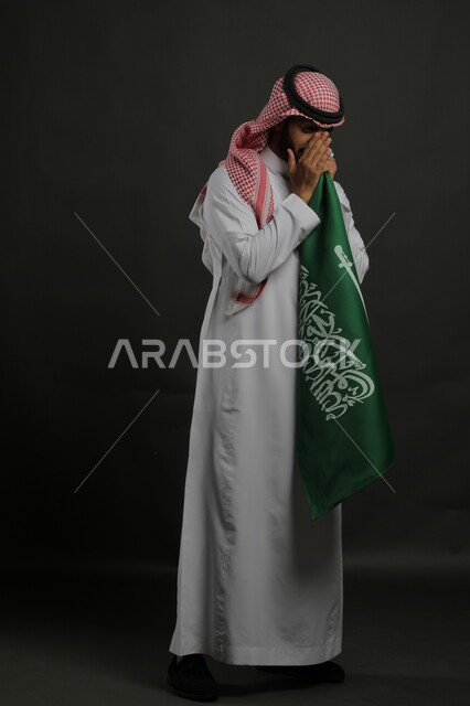 الشعور بالامتنان والفرح في اليوم الوطني السعودي 23 سبتمبر، بورتريه لرجل عربي خليجي سعودي يرتدي الشماغ والثوب التقليدي يحمل بيده علم المملكة ،  يوم العلم  11 مارس ، صورة الجسم بالطول الكامل , خلفية رمادية