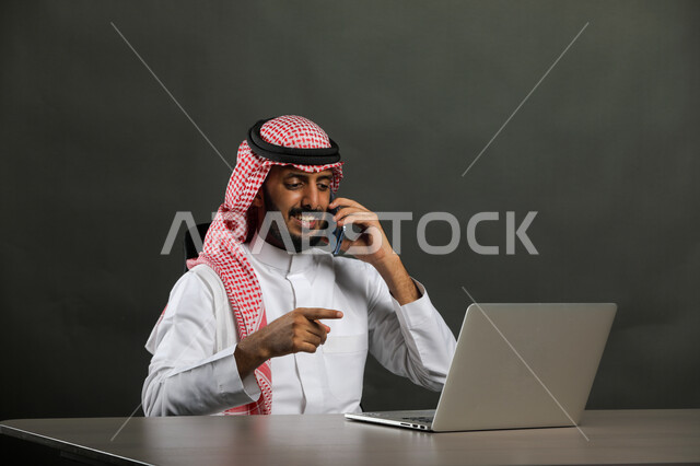 Integrating work with technology and technology, a close-up portrait of a Saudi Gulf Arab man wearing a traditional thobe and shemagh making a phone call via a mobile phone, browsing business management applications via a computer, consultations and inquiries about the mechanism for completing office tasks, black background