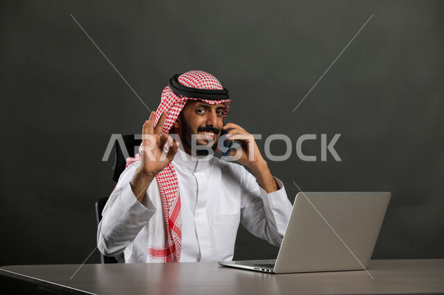 Gestures of quality, satisfaction and acceptance, a close-up portrait of a smiling Saudi Gulf Arab man wearing a traditional thobe and shemagh making a phone call via a mobile phone, working in the office field, completing tasks via a computer, following up on administrative work, black background