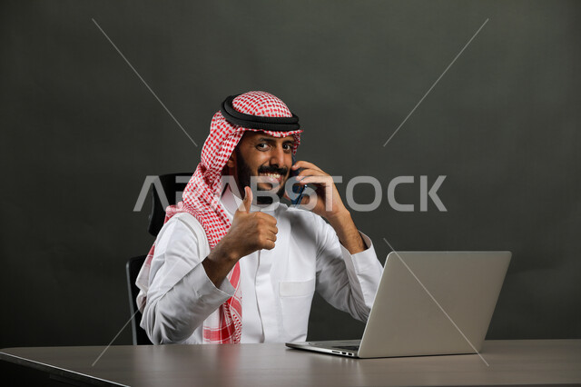 إيماءات الجودة والرضا والقبول, بورتريه مقرب لرجل عربي خليجي سعودي مبتسم يرتدي الثوب التقليدي والشماغ يقوم بإجراء مكالمة هاتفية عبر الهاتف المحمول, العمل في المجال المكتبي, انجاز المهام عبر جهاز الحاسوب, متابعه الأعمال الادارية, خلفية سوداء