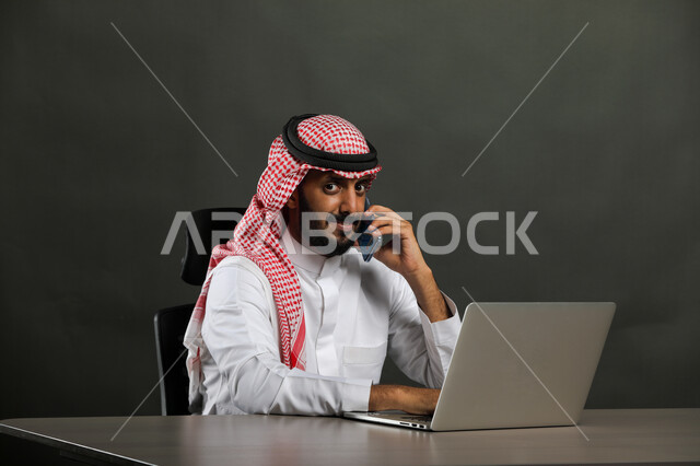 النظر إلى الكاميرا والجلوس في المكتب أمام جهاز الحاسوب, إدارة الأعمال عبر  الأونلاين, بورتريه مقرب لرجل عربي خليجي سعودي يرتدي الثوب التقليدي والشماغ يقوم بإجراء مكالمة هاتفية عبر الهاتف المحمول, البحث عن شواغر وإجراء مقابلات عمل وظيفية, خلفية سوداء