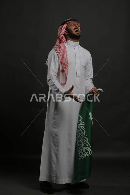 السرور و الحماس أثناء الاحتفال باليوم الوطني السعودي 23 سبتمبر، بورتريه لرجل عربي خليجي سعودي يرتدي الشماغ والثوب التقليدي يحمل بيده علم المملكة ، إيماءات الصراخ من شدة الفرح في يوم العلم  11 مارس ، صورة الجسم بالطول الكامل , خلفية رمادية