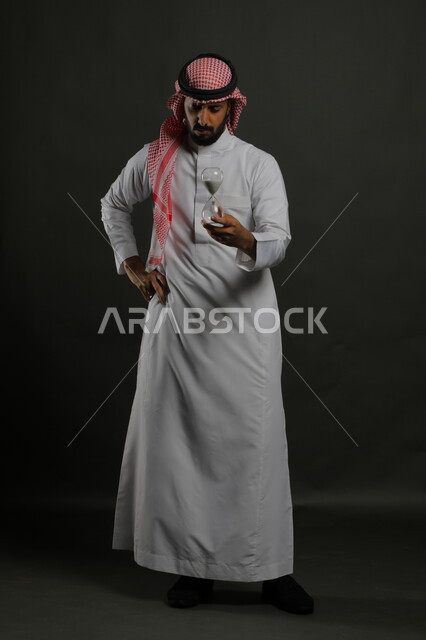 انتظار انقضاء المهلة, مفهوم إدارة الأعمال والتوقيت، بورتريه لشاب عربي سعودي خليجي يرتدي الشماغ والثوب التقليدي يضع يده على خصره يحمل الساعة الرملية الزجاجية (البنكام)، تحديد مواعيد العمل, صورة الجسم بالطول الكامل، خلفية رمادية.