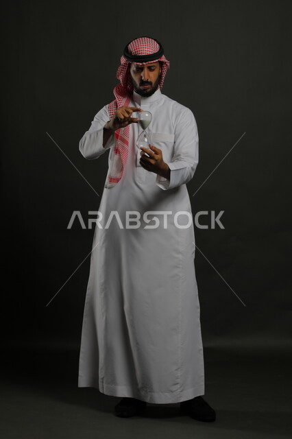 الشعور بالقلق من انقضاء الزمن, مفهوم إدارة الأعمال والتوقيت، بورتريه لرجل عربي سعودي خليجي يرتدي الشماغ والثوب التقليدي يحمل بيده الساعة الرملية الزجاجية (البنكام)، إيماءات الانزعاج والتوتر من نفاذ الوقت، صورة الجسم بالطول الكامل, خلفية رمادية.