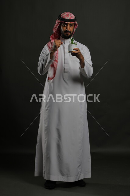 إيماءات النجاح بالعناية بالشتلة, العناية بنباتات الزينة, بورتريه لرجل عربي خليجي سعودي مبتسم يرتدي الثوب التقليدي والشماغ يحمل بيده حوض زراعي صغير, الوقوف باستقامة والنظر إلى الكاميرا بإيماءات السرور والبشاشة, صورة الجسم بالطول الكامل, خلفية رمادية
