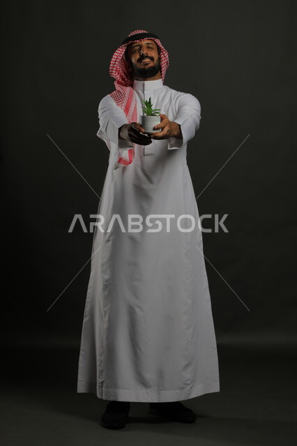 الاهتمام بالشتلات المنزلية, العناية بنباتات الزينة, بورتريه لرجل عربي خليجي سعودي مبتسم يرتدي الثوب التقليدي والشماغ يحمل بيده حوض زراعي صغير, الوقوف باستقامة والنظر إلى الكاميرا بإيماءات السرور والبشاشة, صورة الجسم بالطول الكامل, خلفية رمادية