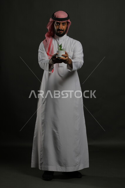 تنقية الهواء  من خلال نباتات الزينة، الاهتمام بالشتلات المنزلية, بورتريه لرجل عربي خليجي سعودي مبتسم يرتدي الثوب التقليدي والشماغ يحمل بيده حوض زراعي صغير, الوقوف باستقامة والنظر إلى الكاميرا بإيماءات السرور والبشاشة, صورة الجسم بالطول الكامل, خلفية رمادية