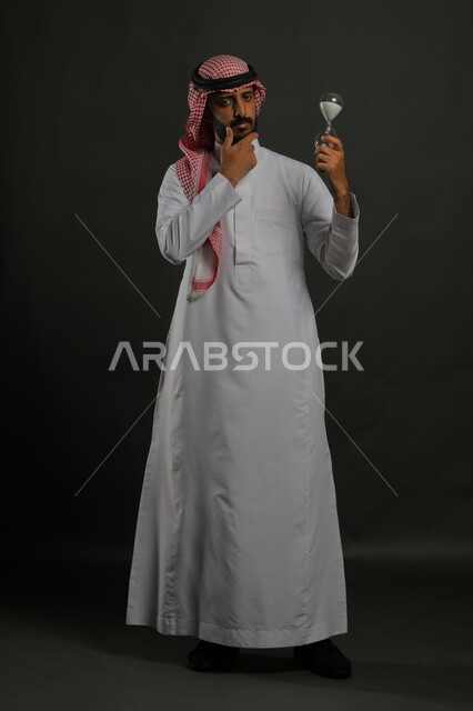  تعزيز السكينة باستخدام الشتلات المنزلية، العناية بنباتات الزينة، بورتريه لرجل عربي خليجي سعودي يرتدي الثوب التقليدي والشماغ يحمل بيده حوض زراعي صغير, الوقوف باستقامة والنظر إلى الكاميرا بايماءات التفكير والاندماج،, صورة الجسم بالطول الكامل, خلفية رمادية