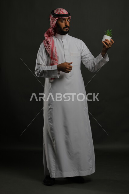  زراعة النباتات الخضراء، الاهتمام بالشتلات المنزلية, بورتريه لرجل عربي خليجي سعودي يرتدي الثوب التقليدي والشماغ يحمل بيده حوض زراعي صغير, الوقوف باستقامة والنظر إلى الكاميرا بايماءات الحيرة والصدمة،  صورة الجسم بالطول الكامل, خلفية رمادية
