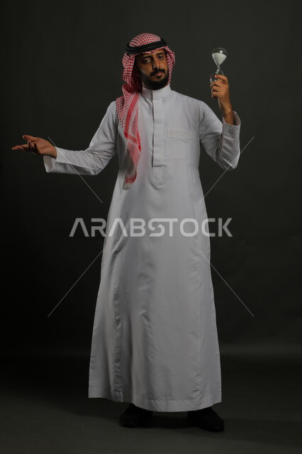 الاهتمام بالشتلات المنزلية، العناية بنباتات الزينة,  بورتريه لرجل عربي خليجي سعودي يرتدي الثوب التقليدي والشماغ يحمل بيده حوض زراعي صغير, الوقوف باستقامة والنظر إلى الكاميرا بإيماءات الحيرة والتردد, صورة الجسم بالكامل, خلفية رمادية