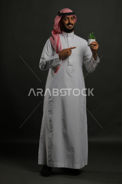 أناقة وجمال نبتات الزينة، الاهتمام بالشتلات المنزلية, بورتريه لرجل عربي خليجي سعودي مبتسم يرتدي الثوب التقليدي والشماغ يشير الى حوض زراعي صغير, الوقوف باستقامة والنظر إلى الكاميرا بإيماءات السرور والبشاشة, صورة الجسم بالطول الكامل, خلفية رمادية
