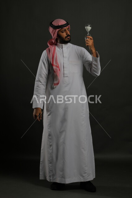 العناية بنباتات الزينة, الاهتمام بالشتلات المنزلية, بورتريه لرجل عربي خليجي سعودي يرتدي الثوب التقليدي والشماغ ينظر الى حوض زراعي صغير, الوقوف باستقامة والنظر إلى الكاميرا بإيماءات التفكير والتركيز, صورة الجسم بالطول الكامل, خلفية رمادية