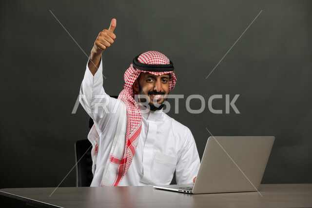 إنجاز المهام عبر جهاز الحاسوب, متابعه الأعمال الادارية, إيماءات الجودة والرضا والقبول, بورتريه مقرب لرجل عربي خليجي سعودي مبتسم يرتدي الثوب التقليدي والشماغ ينظر إلى الكاميرا بسعادة وسرور, العمل في المجال المكتبي, خلفية رمادية