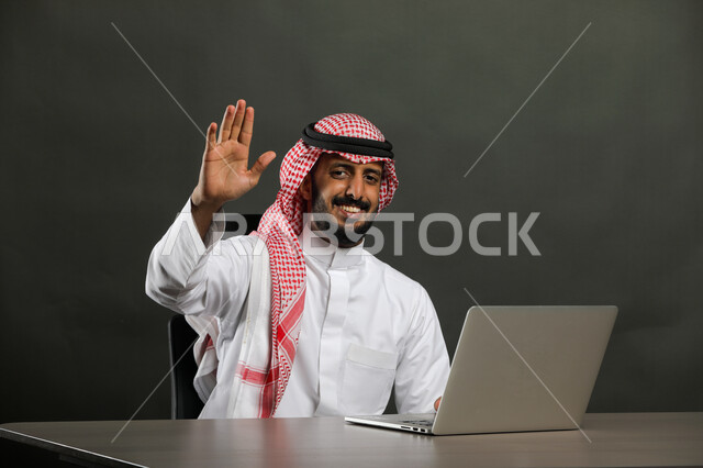 إلقاء التحية والسلام, التعبير عن الترحيب بسرور, بورتريه لرجل عربي خليجي سعودي مبتسم يرتدي الثوب التقليدي والشماغ يرفع كفه للأعلى, إدارة الأعمال المكتبية, الجلوس والعمل عبر جهاز الحاسوب, خلفية رمادية