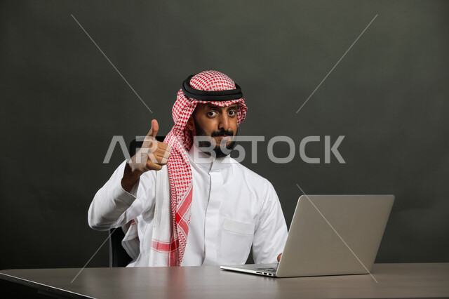 Completing tasks via the computer, following up on administrative work, gestures of quality, satisfaction and acceptance, close-up portrait of a smiling Saudi Gulf Arab man wearing a traditional thobe and shemagh looking at the camera happily and happily, working in the office field, gray background