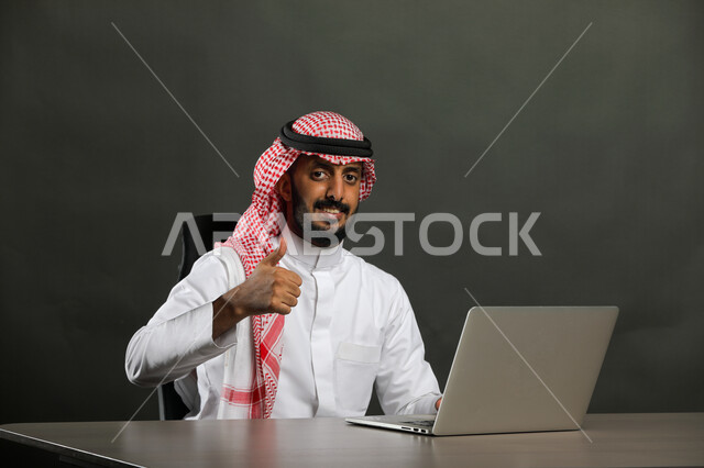متابعه الأعمال الادارية, إيماءات الجودة والرضا والقبول, بورتريه مقرب لرجل عربي خليجي سعودي مبتسم يرتدي الثوب التقليدي والشماغ ينظر إلى الكاميرا بسعادة وسرور, العمل في المجال المكتبي, انجاز المهام عبر جهاز الحاسوب, خلفية رمادية