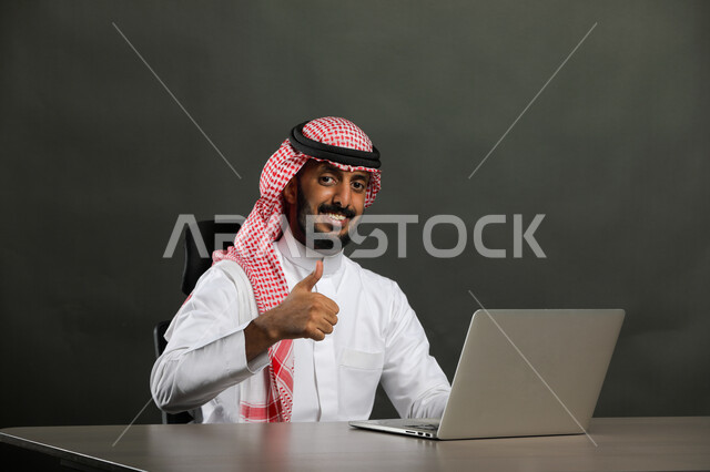إيماءات الجودة والرضا والقبول, بورتريه مقرب لرجل عربي خليجي سعودي مبتسم يرتدي الثوب التقليدي والشماغ ينظر إلى الكاميرا بسعادة وسرور, العمل في المجال المكتبي, انجاز المهام عبر جهاز الحاسوب, متابعه الأعمال الادارية, خلفية رمادية