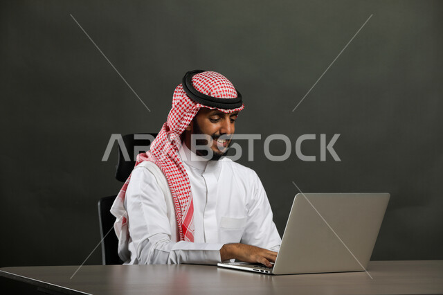 استخدام الأجهزة والتقنيات الحديثة والمتطورة, عقد اجتماع عمل عبر الانترنت, بورتريه مقرب لرجل عربي خليجي سعودي مبتسم يرتدي الثوب التقليدي والشماغ يقوم بتصفح مواقع أخبار السوشال ميديا عبر جهاز الحاسوب, العمل في المجال المكتبي, خلفية رمادية