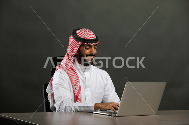 استخدام الأجهزة والتقنيات الحديثة والمتطورة, عقد اجتماع عمل عبر الانترنت, بورتريه مقرب لرجل عربي خليجي سعودي مبتسم يرتدي الثوب التقليدي والشماغ يقوم بتصفح مواقع أخبار السوشال ميديا عبر جهاز الحاسوب, العمل في المجال المكتبي, خلفية رمادية