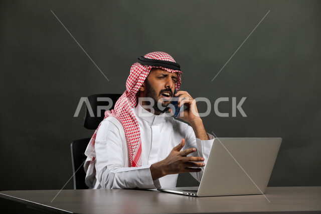  إحتراف وظائف الحاسوب وتكنولوجيا المعلومات، بورتريه مقرب  لرجل عربي خليجي سعودي يرتدي الزي التقليدي والشماغ و يجلس على المكتب أمام اللابتوب ويجري مكالمة عبر الهاتف المحمول، إستخدام الأجهزة التقنية الحديثة، خلفية رمادي
