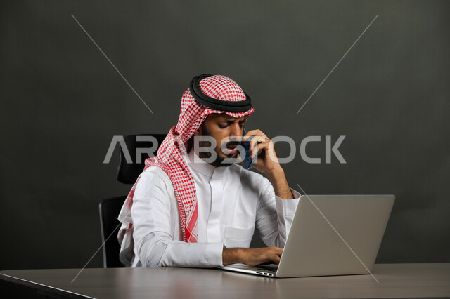 Using modern technologies in remote work, a close-up portrait of a Saudi Gulf Arab man wearing traditional clothing and a shemagh, sitting in front of the laptop and making a call via mobile phone, completing work online through the computer, the ability to pay bills by electronic card, gray background