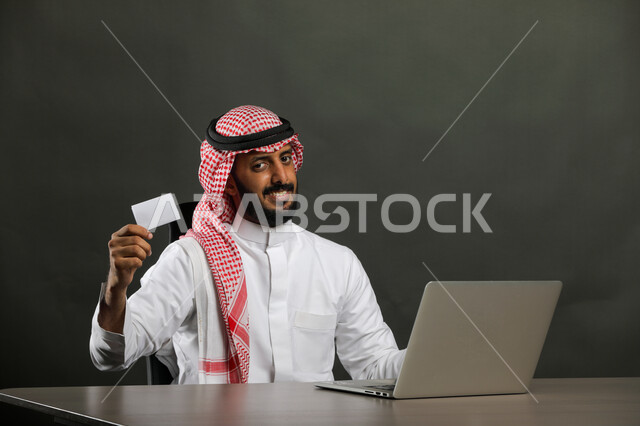 The possibility of paying bills with an electronic card, the development of technology and the use of modern technical devices, a close-up portrait of a Saudi Gulf Arab man wearing a traditional dress and shemagh and holding a blank white card in his hand, the use of modern technologies in remote work, gray background