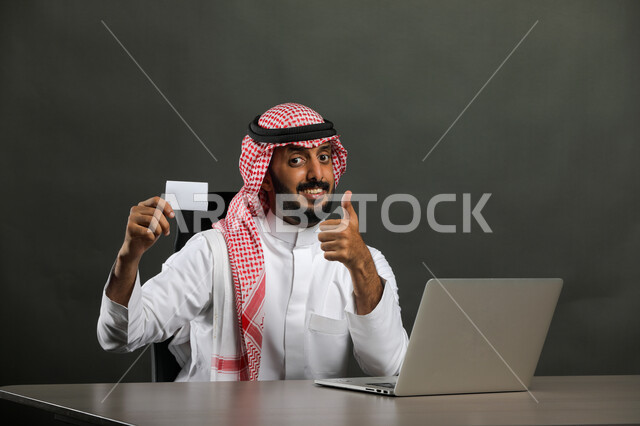  ايماءات الموافقة والاعجاب والقبول، بورتريه مقرب  لرجل عربي خليجي سعودي يرتدي الزي التقليدي والشماغ يجلس على المكتب ويستخدم اللابتوب لإنجاز المهام ، بطاقة بيضاء فارغة، وسائل وتقنيات متطورة وحديثة، الشراء عن طريق بطاقة الائتمان البنكيه، خلفية رمادي