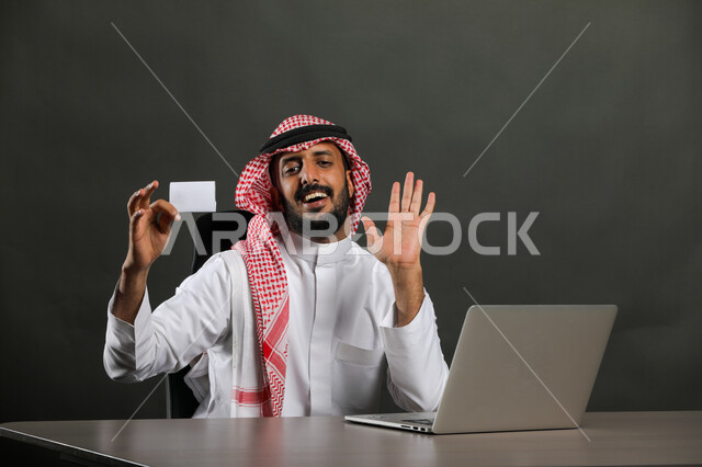 انجاز المهام المكتبية من خلال اللابتوب، بورتريه مقرب  لرجل عربي خليجي سعودي يرتدي الزي التقليدي والشماغ ويقوم بعقد إجتماع عمل عبر الإنترنت وينظر الى بطاقة بيضاء فارغة، توفير خدمة الدفع الالكتروني لطلبات الاونلاين، خلفية رمادي