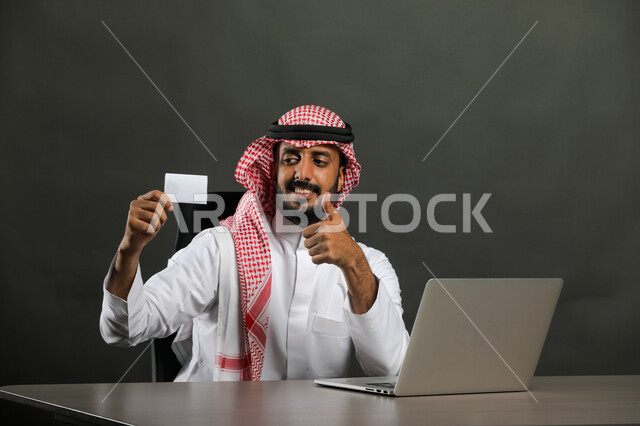  ايماءات الموافقة والاعجاب والقبول، بورتريه مقرب  لرجل عربي خليجي سعودي يرتدي الزي التقليدي والشماغ يجلس على المكتب ويستخدم اللابتوب لإنجاز المهام ، النظر الى بطاقة بيضاء فارغة، وسائل وتقنيات متطورة وحديثة، الشراء عن طريق بطاقة الائتمان البنكيه، خلفية رمادي