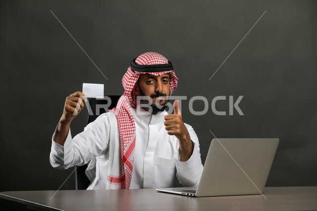 عقد الاجتماع وإدارة المشاريع والأعمال الادارية عبر جهاز الحاسوب, بورتريه مقرب لرجل عربي خليجي سعودي مبتسم يرتدي الثوب التقليدي والشماغ يمسك بطاقة عمل تعريفية, الجلوس والنظر إلى الكاميرا بإيماءات الموافقة والجوده والقبول, خلفية رمادي