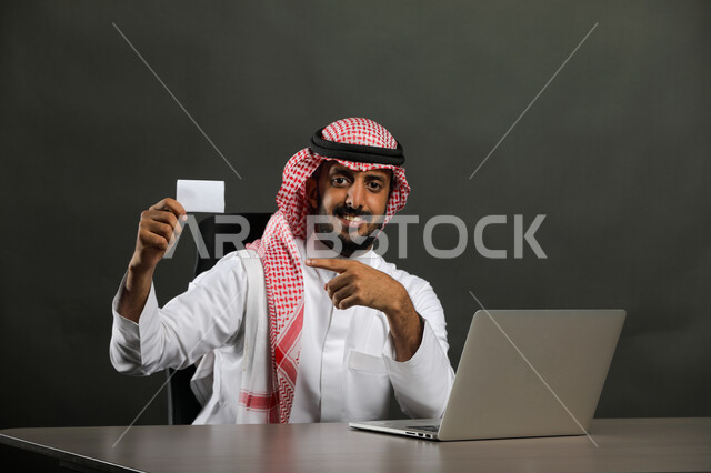 عقد الاجتماع وإدارة المشاريع والأعمال الادارية عبر جهاز الحاسوب, بورتريه مقرب لرجل عربي خليجي سعودي مبتسم يرتدي الثوب التقليدي والشماغ يمسك بطاقة عمل تعريفية, الجلوس والنظر إلى الكاميرا بإيماءات الموافقة والجوده والقبول, خلفية رمادي