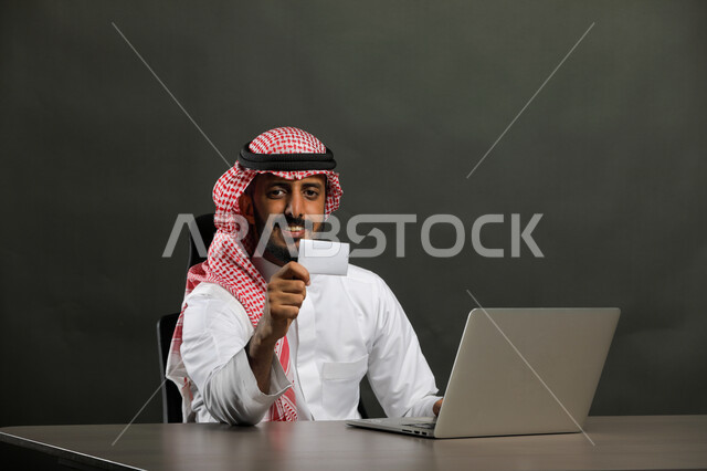 Completing office tasks, project management, and administrative work via the computer, a close-up portrait of a smiling Saudi Gulf Arab man wearing a traditional thobe and shemagh, holding an identification business card, sitting and looking at the camera with happy gestures, gray background