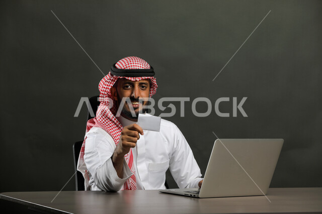 Completing office tasks, project management, and administrative work via the computer, a close-up portrait of a smiling Saudi Gulf Arab man wearing a traditional thobe and shemagh, holding an identification business card, sitting and looking at the camera with happy gestures, gray background