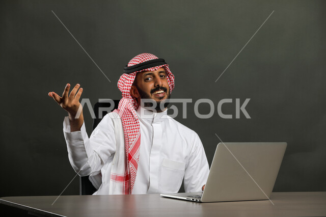 اجتماع ومناقشة عمل عبر الانترنت, مفهوم الحوار والجدال, محاولات الإقناع في موضوع ما, بورتريه مقرب لرجل عربي خليجي سعودي يرتدي الثوب التقليدي والشماغ يقوم باستخدم جهاز الحاسوب ويرفع يديه بإيماءات الاستفسار عن شيء معين, خلفية رمادي