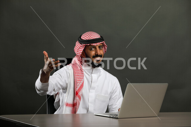 اجتماع ومناقشة عمل عبر الانترنت, مفهوم الحوار والجدال, محاولات الإقناع في موضوع ما, بورتريه مقرب لرجل عربي خليجي سعودي يرتدي الثوب التقليدي والشماغ يقوم باستخدم جهاز الحاسوب ويرفع يديه بإيماءات الاستفسار عن شيء معين, خلفية رمادي