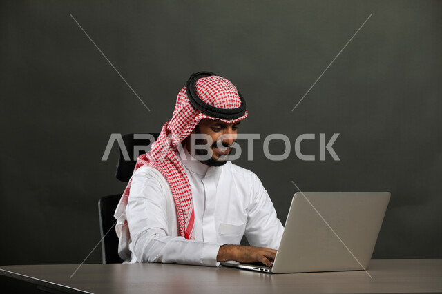 استخدام الأجهزة والتقنيات الحديثة والمتطورة, عقد اجتماع عمل عبر الانترنت, بورتريه مقرب لرجل عربي خليجي سعودي مبتسم يرتدي الثوب التقليدي والشماغ يقوم بتصفح مواقع أخبار السوشال ميديا عبر جهاز الحاسوب, العمل في المجال المكتبي, خلفية رمادية