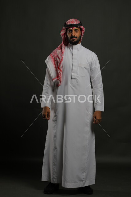 مفهوم الرجولة و الاهتمام بالمظهر الخارجي، الوقوف باستقامة وانضباط، الثقة بالنفس والشعور بالقوة، بورتريه لشاب عربي خليجي سعودي مبتسم يرتدي الشماغ والثوب التقليدي يضع يديه على جانبه و ينظر إلى الكاميرا بإيماءات السرور، صورة الجسم بالطول الكامل، خلفية رمادية.