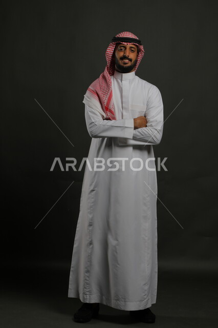 الاهتمام بالمظهر الخارجي، مفهوم الاناقه و الرجولة ،الوقوف باستقامة وثقه بالنفس، بورتريه لشاب وسيم عربي خليجي سعودي مسرور يرتدي الشماغ و الزي التقليدي ينظر إلى الكاميرا بإيماءات أيدي متقاطعه، صورة الجسم بالطول الكامل، خلفية رمادية